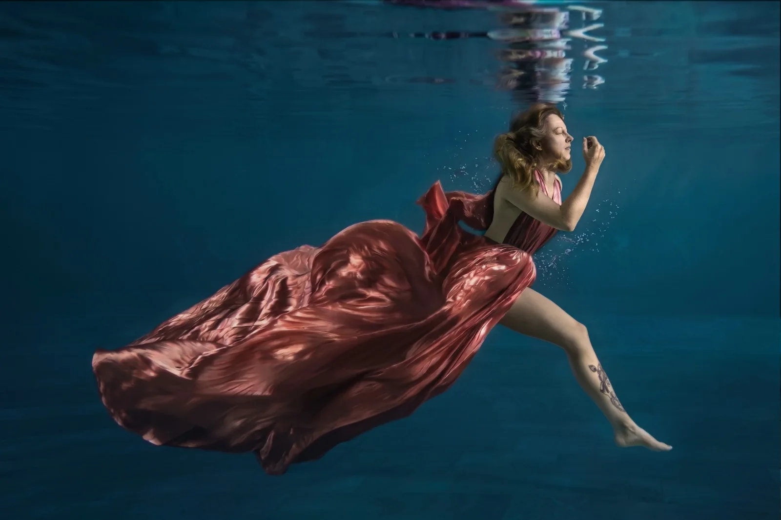 Frau im fliessenden roséroten Satinkleid gleitet mit geschlossenen Augen durch tiefblaues Wasser – Beyond Gravity, Unterwasser-Portrait