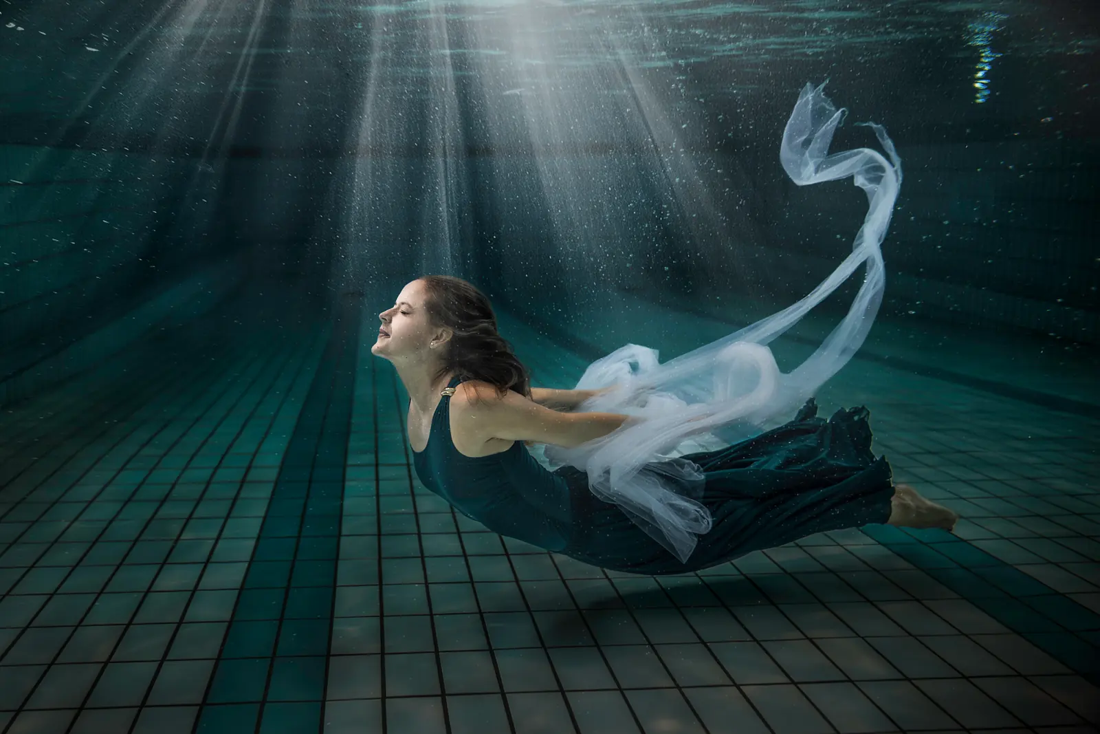 Frau im dunklen Kleid schwebt mit weissem Tuch unter Wasser im Lichtstrahl