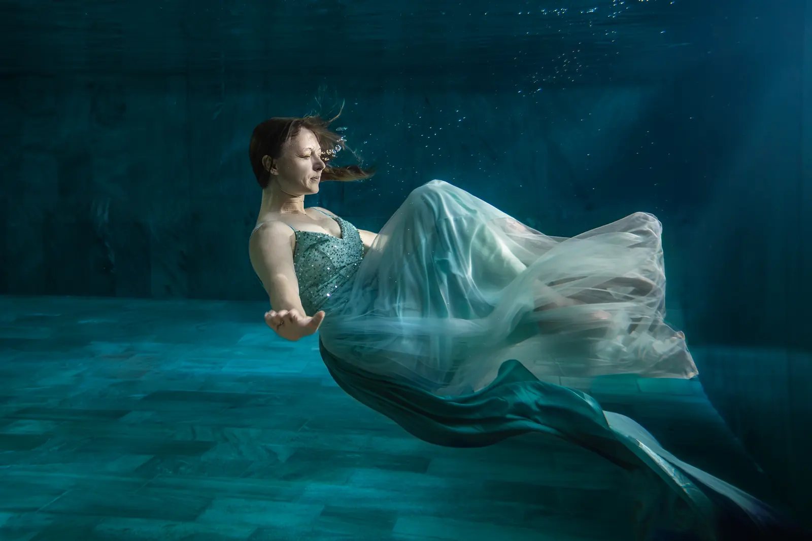 Frau im mintfarbenen Kleid schwebt rücklings unter Wasser, weicher Lichtstrahl von oben
