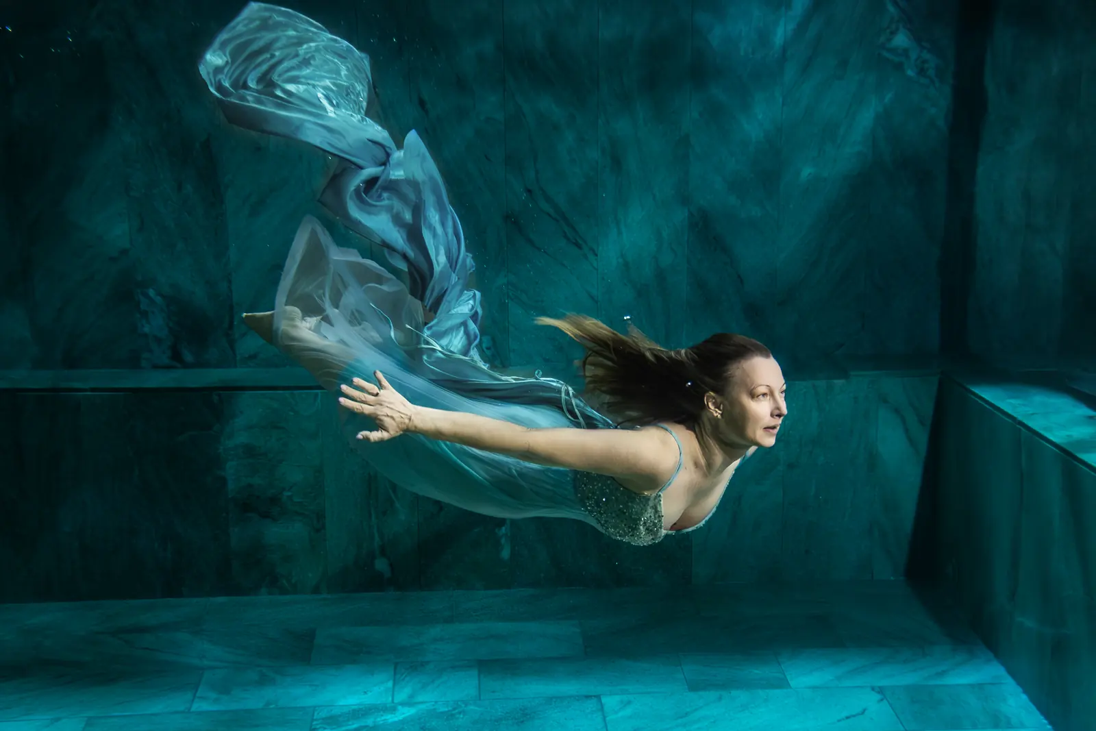 Frau im hellen Kleid taucht unter Wasser nach vorn, Stoff und Haare ziehen hinter ihr her