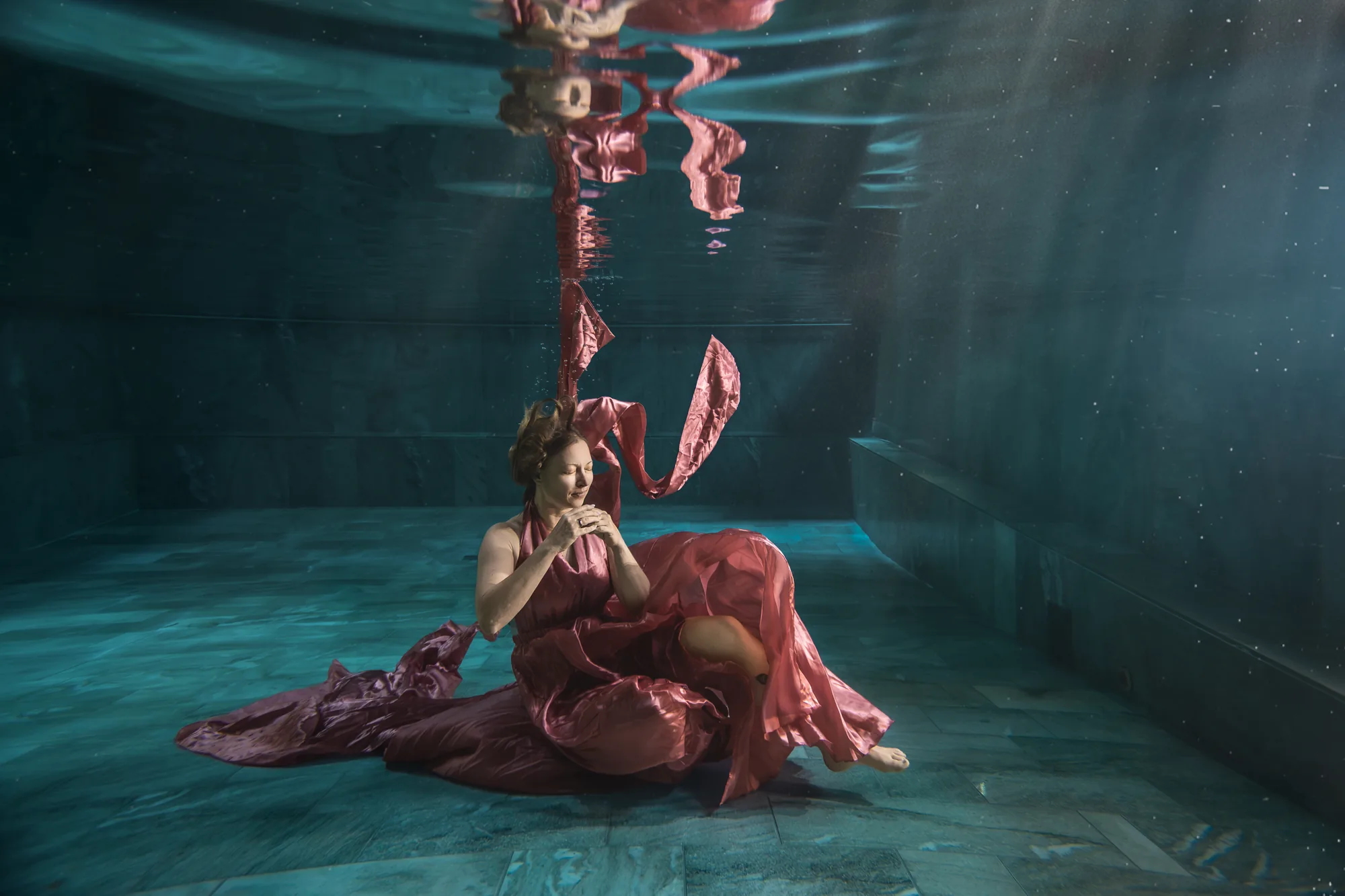 Frau im roséfarbenen Satinkleid sitzt mit geschlossenen Augen am Poolboden, der Stoff spiegelt sich an der Wasseroberfläche – Unterwasser-Portrait