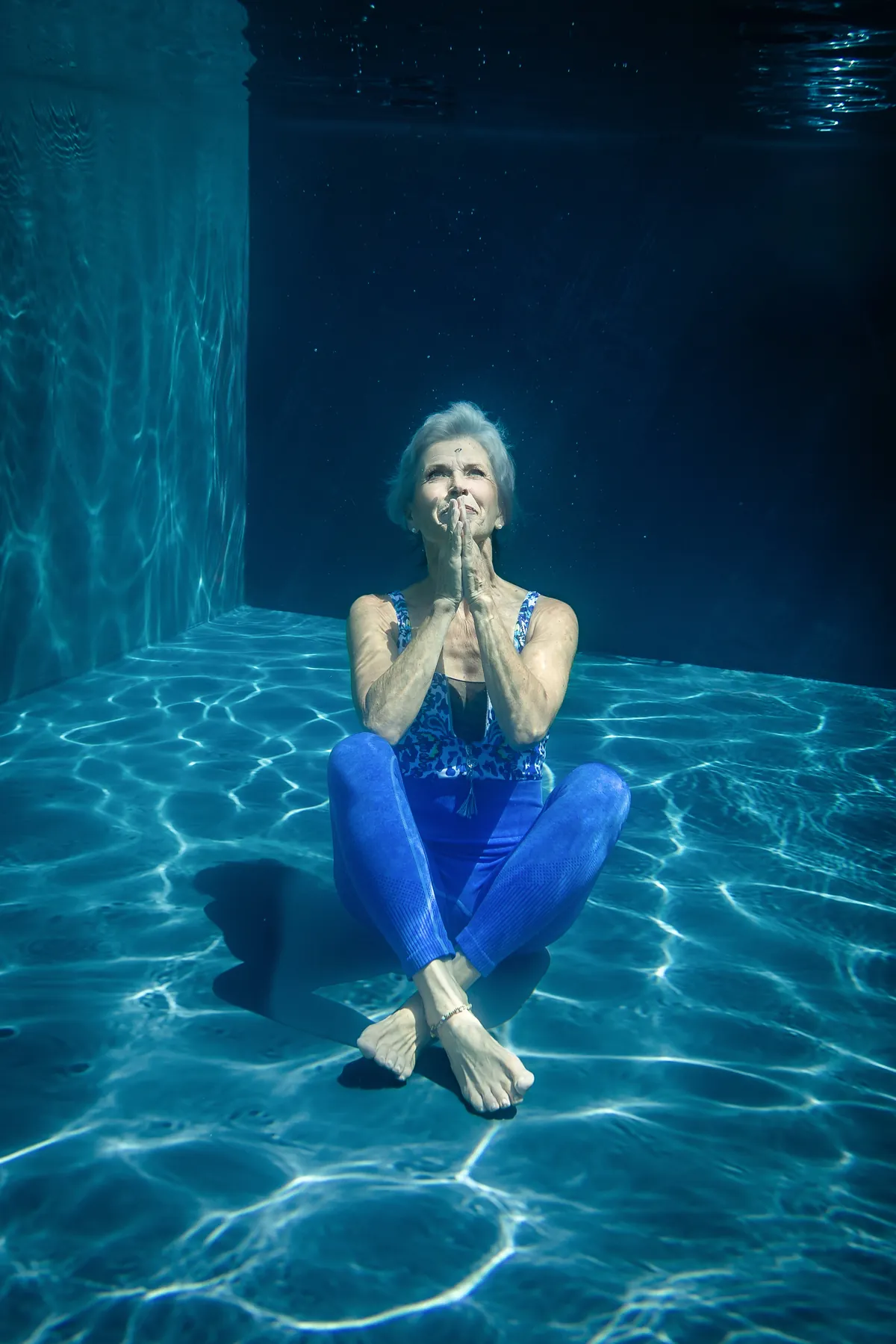 Frau sitzt mit gefalteten Händen im Lotussitz auf dem Poolboden, Lichtreflexe im blauen Wasser – Unterwasser-Portrait von Stefanie Blochwitz